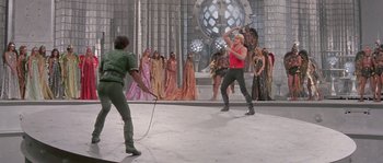 Movie still from “Flash Gordon” (1980), directed by Mike Hodges – A man and a woman are playing a game with a rope in front of a group of people; Wide shot, Over the shoulder angle