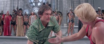 Movie still from “Flash Gordon” (1980), directed by Mike Hodges – A man in a green shirt is talking to another man; Medium shot, Over the shoulder angle