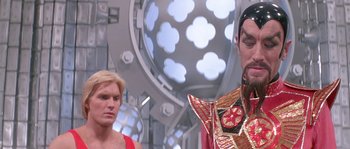 Movie still from “Flash Gordon” (1980), directed by Mike Hodges – A man in a red shirt is standing next to another man in a red shirt; Close Up shot, Over the shoulder angle