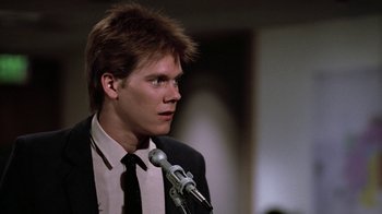 Movie still from “Footloose” (1984), directed by Herbert Ross – A man in a suit and tie is talking into a microphone; Close Up shot, Over the shoulder angle
