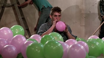 Movie still from “Footloose” (1984), directed by Herbert Ross – A young man blowing up a balloon in front of a bunch of green balloons; Medium shot, Low angle