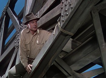 Movie still from “For Whom the Bell Tolls” (1943), directed by Sam Wood – A man in a hat and a brown shirt is climbing a wooden structure; Medium shot, Low angle