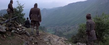 Movie still from “Force 10 from Navarone” (1978), directed by Guy Hamilton – A man standing on top of a mountain looking at a bridge; Wide shot, Low angle