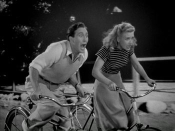 Movie still from “Four Daughters” (1938), directed by Michael Curtiz – A man and a woman riding bicycles on a field; Medium shot, High angle