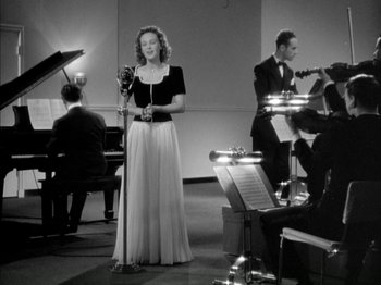 Movie still from “Four Daughters” (1938), directed by Michael Curtiz – A black and white photo of a woman holding a microphone; Wide shot, Low angle