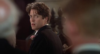 Movie still from “Four Weddings and a Funeral” (1994), directed by Mike Newell – A man in a black suit and a pink flower in his hair; Close Up shot, Over the shoulder angle