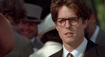 Movie still from “Four Weddings and a Funeral” (1994), directed by Mike Newell – A man in a suit and tie wearing glasses; Close Up shot, Over the shoulder angle