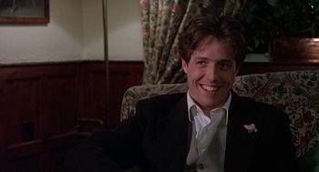 Movie still from “Four Weddings and a Funeral” (1994), directed by Mike Newell – A man sitting in a chair smiling for the camera; Close Up shot, Low angle