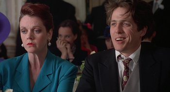 Movie still from “Four Weddings and a Funeral” (1994), directed by Mike Newell – A man and a woman sitting next to each other in front of other people; Close Up shot, Low angle