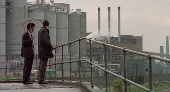 Movie still from “Four Weddings and a Funeral” (1994), directed by Mike Newell – A man standing on the side of a road next to a railing; Wide shot, Low angle