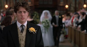 Movie still from “Four Weddings and a Funeral” (1994), directed by Mike Newell – A man in a suit and tie standing in front of a bride and groom; Close Up shot, Over the shoulder angle
