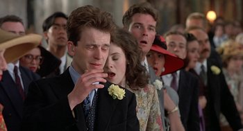Movie still from “Four Weddings and a Funeral” (1994), directed by Mike Newell – A man smoking a cigarette in front of a group of people; Close Up shot, Over the shoulder angle
