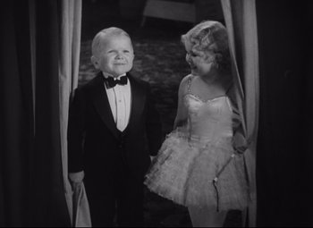 Movie still from “Freaks” (1932), directed by Tod Browning – A little boy in a tuxedo standing next to a woman in a white dress; Medium shot, High angle