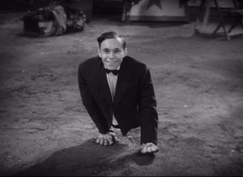 Movie still from “Freaks” (1932), directed by Tod Browning – A black and white photo of a man in a suit and bow tie kneeling on the ground; Medium shot, High angle