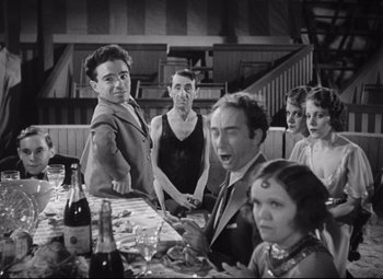 Movie still from “Freaks” (1932), directed by Tod Browning – A group of people sitting at a table with drinks; Medium shot, Over the shoulder angle