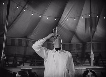 Movie still from “Freaks” (1932), directed by Tod Browning – A man standing in front of an awning holding a drink; Medium shot, Low angle