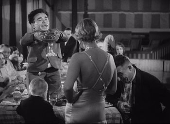 Movie still from “Freaks” (1932), directed by Tod Browning – A man standing next to a group of people at a table; Medium shot, High angle