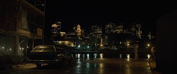 Movie still from “Free Fire” (2016), directed by Ben Wheatley – Cars parked in a parking lot in front of a city skyline at night; Extreme Wide shot, High angle