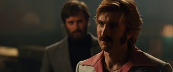 Movie still from “Free Fire” (2016), directed by Ben Wheatley – A man with a mustache and a beard standing next to another man with a beard; Close Up shot, Over the shoulder angle