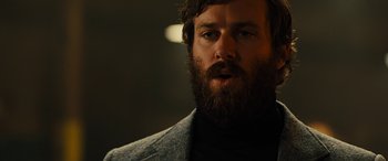 Movie still from “Free Fire” (2016), directed by Ben Wheatley – A man with a beard and a jacket on; Close Up shot, Over the shoulder angle
