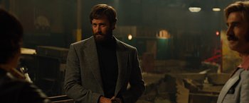 Movie still from “Free Fire” (2016), directed by Ben Wheatley – A man with a beard wearing a suit and tie; Medium shot, Over the shoulder angle