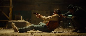 Movie still from “Free Fire” (2016), directed by Ben Wheatley – A man sitting on the ground holding a gun; Wide shot, Low angle