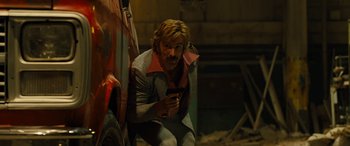 Movie still from “Free Fire” (2016), directed by Ben Wheatley – A man crouching down next to a red truck; Medium shot, Low angle
