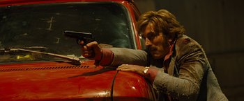 Movie still from “Free Fire” (2016), directed by Ben Wheatley – A man holding a gun while standing next to a red truck; Medium shot, Low angle