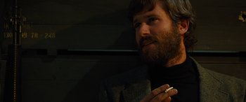 Movie still from “Free Fire” (2016), directed by Ben Wheatley – A man with a beard smoking a cigarette in the dark; Close Up shot, Over the shoulder angle