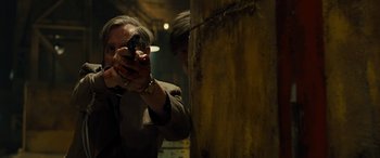 Movie still from “Free Fire” (2016), directed by Ben Wheatley – A man holding a gun while standing next to a wall; Medium shot, Over the shoulder angle