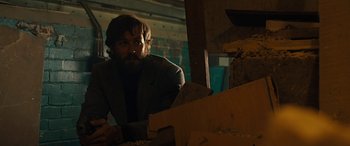 Movie still from “Free Fire” (2016), directed by Ben Wheatley – A man in a suit and beard looking at something in a box; Medium shot, Low angle