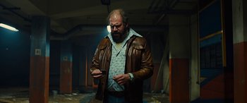 Movie still from “Free Fire” (2016), directed by Ben Wheatley – A man wearing a brown leather jacket holding a cigarette; Medium shot, Low angle