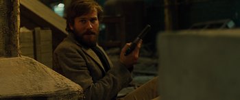 Movie still from “Free Fire” (2016), directed by Ben Wheatley – A man sitting in a chair holding a remote control; Medium shot, Over the shoulder angle