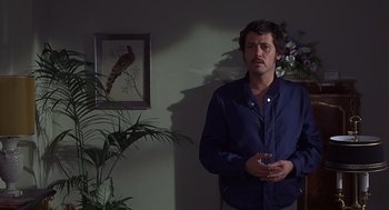 Movie still from “Frenzy” (1972), directed by Alfred Hitchcock – A man standing next to a plant in a living room; Medium shot, Over the shoulder angle