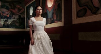 Movie still from “Frida” (2002), directed by Julie Taymor – A woman in a white dress standing in front of a painting; Medium shot, Low angle