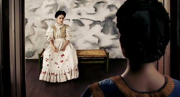 Movie still from “Frida” (2002), directed by Julie Taymor – A painting of a woman sitting on top of a bench next to another woman; Medium shot, Overhead angle