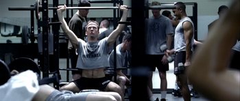 Movie still from “Friday Night Lights” (2004), directed by Peter Berg – A man that is lifting a barbell in a gym; Medium shot, Low angle