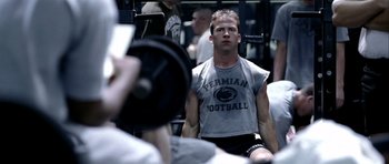 Movie still from “Friday Night Lights” (2004), directed by Peter Berg – A man sitting on a bench in front of a group of people; Medium shot, Over the shoulder angle