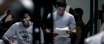 Movie still from “Friday Night Lights” (2004), directed by Peter Berg – A young man reading a book in front of a group of people; Medium shot, Over the shoulder angle