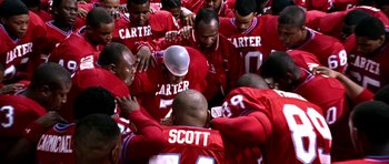 Movie still from “Friday Night Lights” (2004), directed by Peter Berg – A group of football players huddle together in a huddle; Medium shot, Low angle