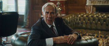 Movie still from “From Paris with Love” (2010), directed by Pierre Morel – An older man sitting in a chair in front of a wooden wall; Close Up shot, Over the shoulder angle