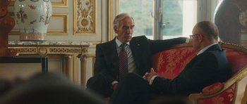 Movie still from “From Paris with Love” (2010), directed by Pierre Morel – An older man wearing a suit and tie sitting on a chair; Medium shot, Over the shoulder angle
