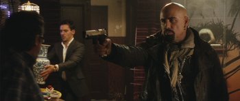 Movie still from “From Paris with Love” (2010), directed by Pierre Morel – A man holding a gun in front of another man in a room; Medium shot, Over the shoulder angle