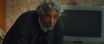 Movie still from “From Paris with Love” (2010), directed by Pierre Morel – An older man with grey curly hair and wearing a sweater; Close Up shot, Over the shoulder angle