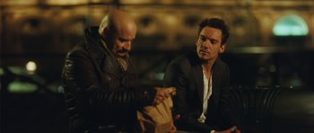 Movie still from “From Paris with Love” (2010), directed by Pierre Morel – Two men sitting at a table with a bag of food in front of them; Medium shot, Over the shoulder angle