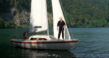Movie still from “Funny Games” (1997), directed by Michael Haneke – A man standing on the front of a sail boat in a body of water; Wide shot, Low angle