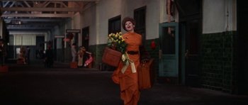 Movie still from “Funny Girl” (1968), directed by William Wyler – A woman dressed in orange carrying a bunch of yellow flowers; Wide shot, Low angle