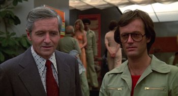Movie still from “Futureworld” (1976), directed by Richard T. Heffron – Two men standing next to each other in front of a group of people; Close Up shot, Over the shoulder angle