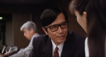 Movie still from “Gamera 3: Revenge of Iris” (1999), directed by Shûsuke Kaneko – A man in a suit and glasses sitting at a table with other people; Close Up shot, Over the shoulder angle