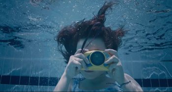 Movie still from “Aftersun” (2022), directed by Charlotte Wells – A woman holding up a camera underwater in a pool; Extreme Close Up shot, Overhead angle